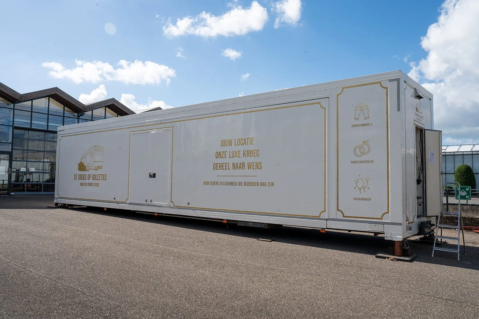 De Kroeg XL van buiten: een witte trailer met gouden belettering, opgesteld op een plein.