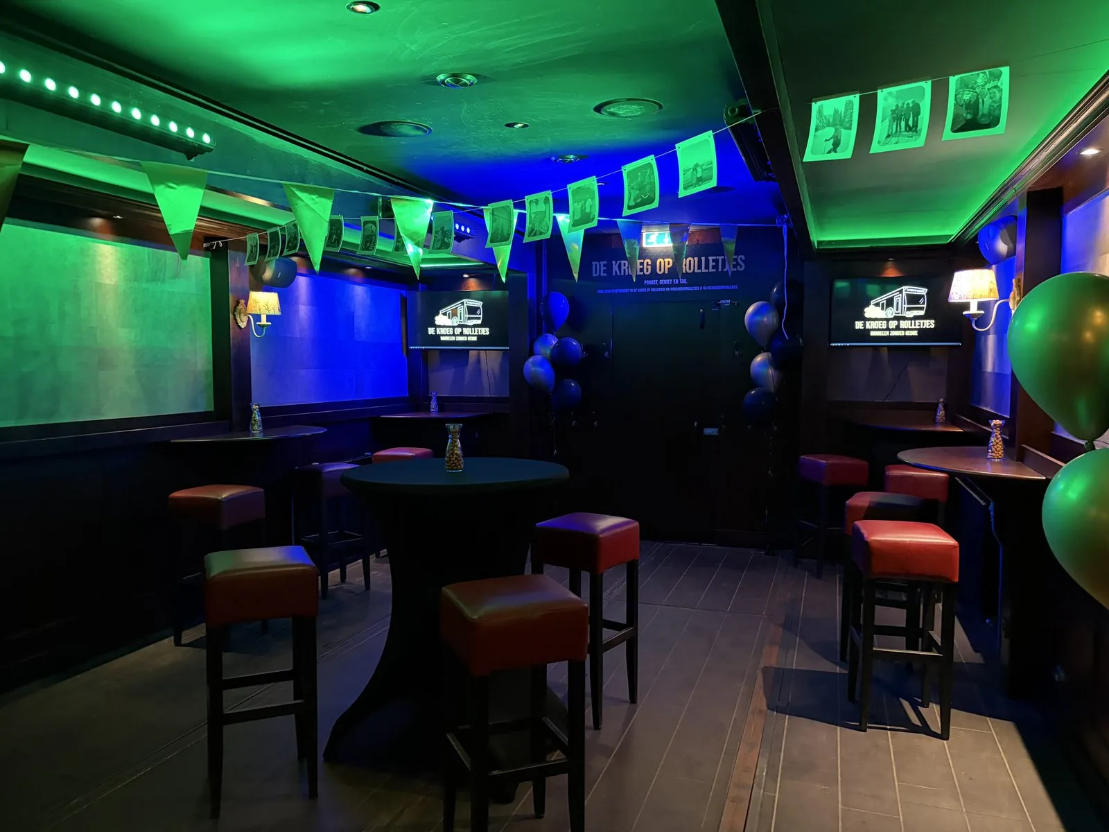 De Kroeg XL in groen en blauw licht, met vlaggetjes en foto’s aan een lijn.